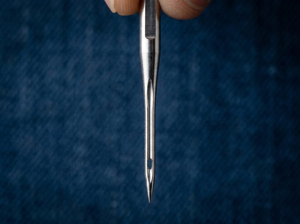 close-up of a flat-shank denim sewing machine needle showing the reinforced blade thickness and scarf, with the eye positioned just above the sharp point