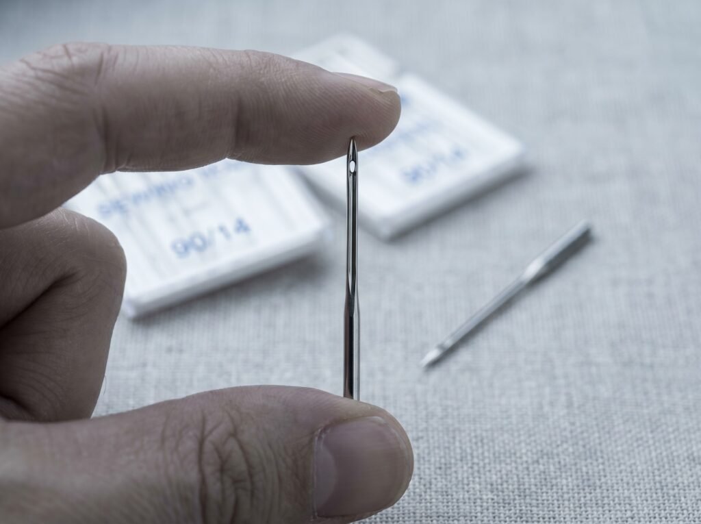 Close-up of a 130/705 H sewing machine needle showing the eye and blade; the flat shank sits at the top (held in the clamp) and the scarf faces away